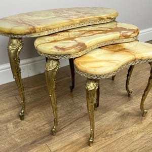 May include: A set of three nesting tables. Each table has an oval onyx top with brown and cream veining. The tables have ornate, gold-coloured metal legs and trim. The tables are arranged in a nested configuration, showcasing a vintage design.