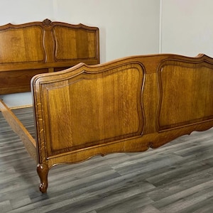 May include: A wooden bed frame featuring a tall headboard and a matching footboard, both constructed with medium-brown oak wood. The headboard and footboard display curved, arched top edges, raised panel detailing, and carved cabriole legs.