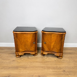 Puede incluir: Un par de mesitas de noche de madera con un acabado de veta de madera marrón claro, una superficie superior negra lisa y una sola puerta en cada unidad. Cada mueble se apoya sobre cuatro patas curvas e incluye un pequeño pomo metálico redondo.