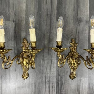 Puede incluir: Dos apliques de pared dorados y ornamentados, cada uno con dos bombillas en forma de vela. Los apliques tienen un diseño floral detallado y están montados en una pared de madera gris.