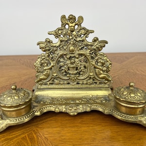 May include: Ornate antique brass inkwell set with two lidded ink pots and a decorative central support featuring cherubs and floral designs. The inkwell set is made of brass and has an aged patina.