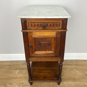 May include: Antique wooden bedside cabinet with a marble top. The cabinet has a small drawer and a cabinet door with a decorative handle. The bottom has a shelf for storage. The cabinet is 76 cm tall.