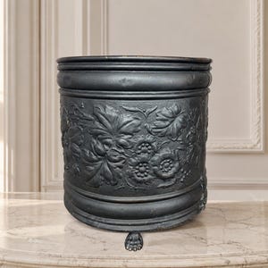 May include: A cylindrical black metal planter featuring a raised relief pattern of leaves and flowers around the exterior. The vessel has a smooth rim at the top and is supported by three small, detailed claw-shaped feet at the base.