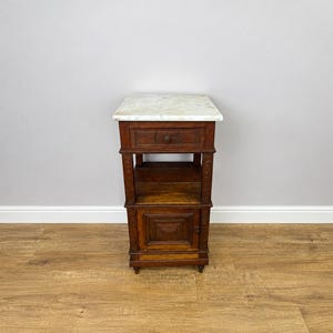 May include: A dark brown wooden bedside table featuring a white marble top, a single upper drawer with a small round knob, an open middle shelf, and a lower cabinet door with a square recessed panel design, supported by four small feet.