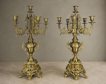 May include: A pair of matching gold-toned metal candelabras, each featuring five candle holders arranged on curved arms. The central stem rises from a decorative urn-shaped base, supported by a square pedestal with ornate relief detailing.