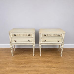 May include: A pair of off-white bedside tables, each featuring two drawers with dark circular knobs. The tables have a rectangular top, fluted detailing on the upper drawer front, and four slender, tapered legs with carved accents at the top.