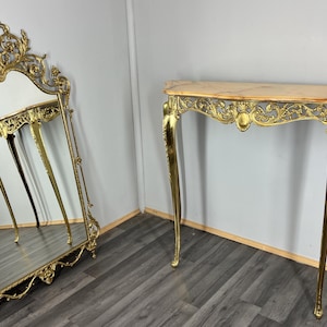 May include: A gold-coloured, ornate console table with a marble top and matching mirror. The table has curved legs and intricate detailing. The mirror is tall and features a decorative frame, reflecting the table. The style is vintage.