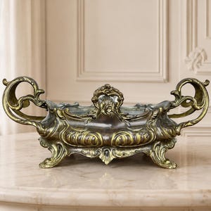 Op de afbeelding: Een versierde, rechthoekige metalen schaal met een donkerbronzen afwerking en goudkleurige accenten. De schaal is voorzien van twee grote, gekrulde handvatten aan de zijkanten, een centraal decoratief ornament en vier kleine, gebogen voetjes.
