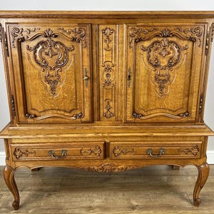 May include: An antique wooden cabinet with elaborate carvings. The cabinet has two doors at the top, decorated with floral and scrollwork, and two drawers below. The wood has a warm, golden-brown finish, and the cabinet stands on curved legs.
