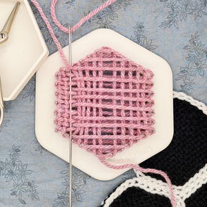 May include: A pink yarn project in progress, with a needle and thread. The project is on a white hexagonal base. A pair of decorative scissors and a white tray are also visible. The background is a blue floral pattern.