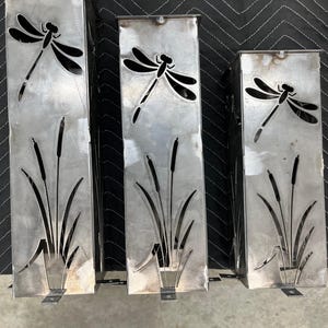May include: Three rectangular metal garden art pieces with cut-out dragonfly and cattail designs. The metal has a brushed finish. Each piece has a small, rectangular base. The dragonflies are black and the cattails are cut out of the metal.