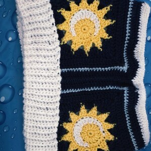 May include: Navy blue and white crocheted blanket with two yellow sun and moon motifs. The blanket has a white border and a ribbed white edge. The sun and moon design is in the center of the blanket.