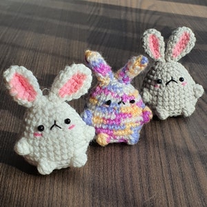 Pode incluir: Três peluches de coelho de crochê. Um é branco com olhos pretos, outro é cinza com orelhas cor-de-rosa e o último é multicolorido com fio rosa, amarelo e roxo.