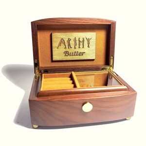 Puede incluir: Una caja de m&uacute;sica de madera con una tapa con bisagras y un pomo de lat&oacute;n. La tapa est&aacute; abierta y revela un peque&ntilde;o compartimento con un forro de terciopelo. La caja de m&uacute;sica est&aacute; grabada con las palabras "Army Butler".