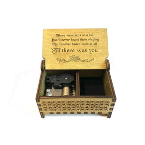 Puede incluir: Caja de m&uacute;sica de madera con un dise&ntilde;o tallado y un mecanismo musical negro. La tapa est&aacute; abierta y presenta una cita: "There were bells on a hill But I never heard them ringing No, I never heard them at all 'Til there was you".