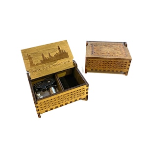 May include: A wooden music box with a carved design on the lid. The lid is open and shows a skyline of New York City with the text "New York New York". The music box is made of dark wood and has a black velvet interior.