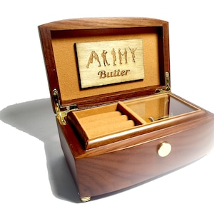 Puede incluir: Una caja de m&uacute;sica de madera con una tapa transparente y un interior de terciopelo marr&oacute;n. La caja tiene un peque&ntilde;o compartimento para joyas y un mecanismo de cuerda. La tapa tiene una placa personalizada con el texto "ARMY Butter".