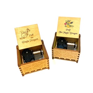 May include: Two wooden music boxes with a dragon and a flower design. The boxes are open and the music mechanisms are visible. The text on the boxes reads "Puff the Magic Dragon".