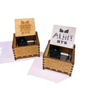 May include: Two wooden music boxes with intricate designs. The lids are open, revealing the inner workings of the music boxes. The top of each box features a different design. One box has a silhouette of seven figures with the text "BTS" below. The other box has the text "ARMY BTS" on the top.