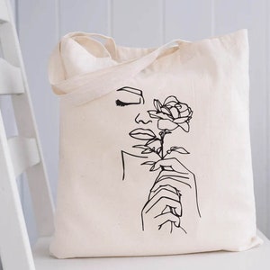 May include: A white tote bag with a black line drawing of a woman's face and a rose. The woman is holding the rose in her hand.