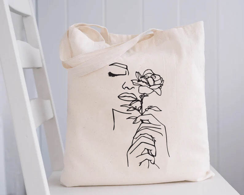 May include: A white tote bag with a black line drawing of a woman's face and a rose. The woman is holding the rose in her hand.