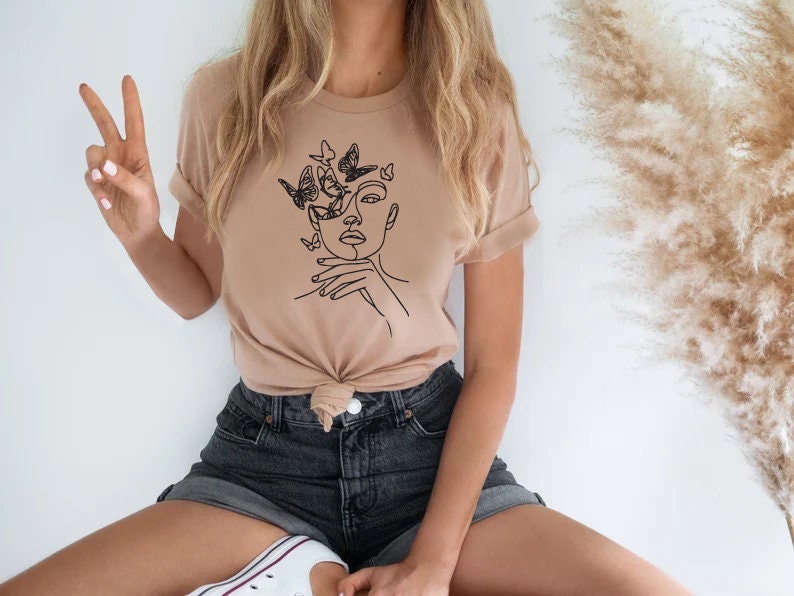 May include: Tan-colored t-shirt featuring a minimalist line art design of a face with butterflies. The black graphic is on the front of the shirt. The sleeves are rolled up. The person is wearing black denim shorts and white sneakers.