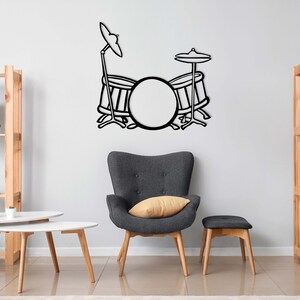 Op de afbeelding: Zwart-witte silhouet van een drumstel op een witte muur. Het drumstel bestaat uit een basdrum, twee kleinere drums en twee bekkens. Het drumstel staat in een woonkamer met een grijze fauteuil, een houten salontafel en twee houten planken.