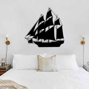 May include: A black metal silhouette of a ship hangs above a white bed with white bedding and a beige throw blanket. Two bedside tables with wooden tops and drawers are on either side of the bed. Each table has a lamp with a gold base and a white bulb.