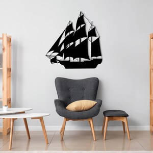 May include: Black metal silhouette of a sailboat mounted on a white wall in a living room setting. The sailboat is centered on the wall and is surrounded by furniture, including a gray armchair, a wooden shelf, and a white coffee table.
