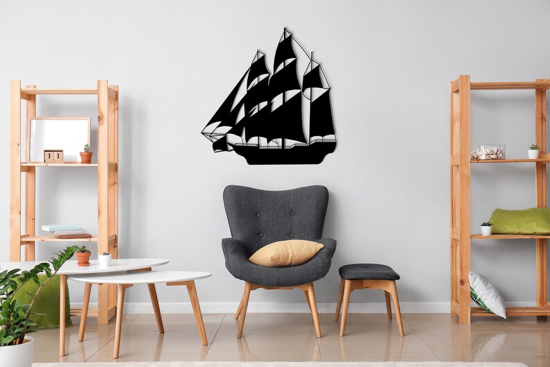 May include: Black metal silhouette of a sailboat mounted on a white wall in a living room setting. The sailboat is centered on the wall and is surrounded by furniture, including a gray armchair, a wooden shelf, and a white coffee table.