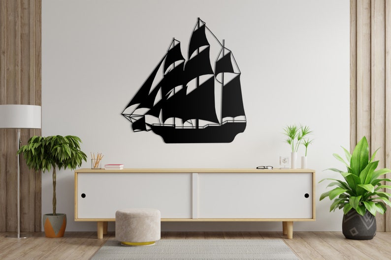 May include: Black metal silhouette of a sailboat with multiple sails, mounted on a white wall above a white dresser with a light wood top. The dresser is in a room with wood floors and two potted plants.
