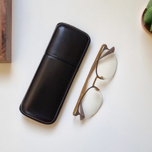 smooth leather eyeglasses case in black on a white table. Shown closed with eyeglasses next to it to show the proportion between the eyeglasses and the leather case.