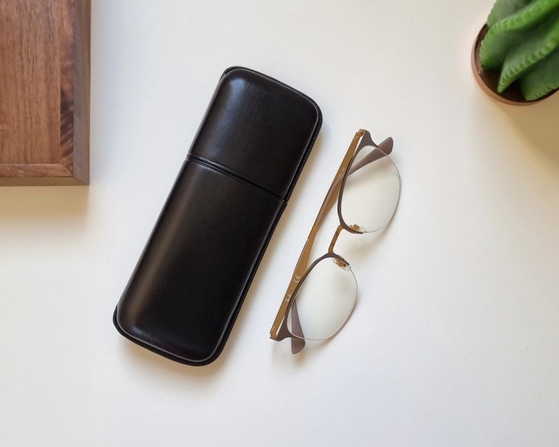 smooth leather eyeglasses case in black on a white table. Shown closed with eyeglasses next to it to show the proportion between the eyeglasses and the leather case.