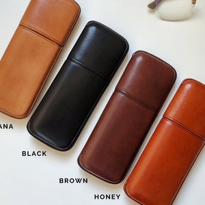 May include: Four leather eyeglass cases in different colors: Havana, Black, Brown, and Honey. The cases are lined up horizontally on a white surface.