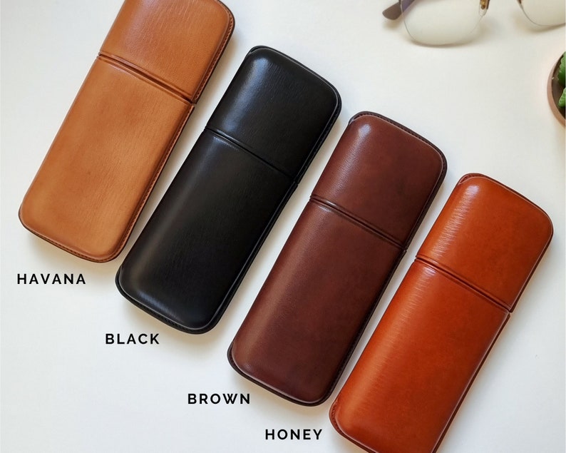 May include: Four leather eyeglass cases in different colors: Havana, Black, Brown, and Honey. The cases are lined up horizontally on a white surface.