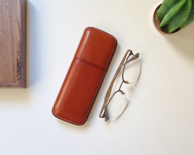 May include: A brown leather glasses case with a textured finish. The case is closed and lying on a white surface.