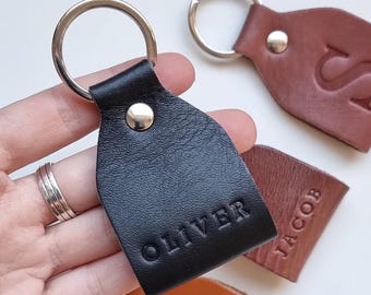 May include: Black leather keychain with the name "OLIVER" embossed. Other keychains in brown and tan leather are visible, with names and initials embossed. Each keychain has a silver metal ring and a metal rivet.