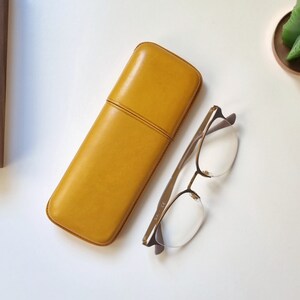 May include: A yellow leather glasses case with a subtle seam down the center. The case is lying on a white surface next to a pair of clear-framed glasses with gold accents.