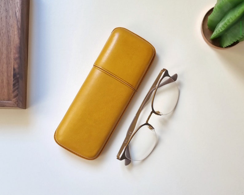 May include: A yellow leather glasses case with a subtle seam down the center. The case is lying on a white surface next to a pair of clear-framed glasses with gold accents.