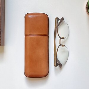 May include: A brown leather glasses case with a gold metal frame glasses case with clear lenses.