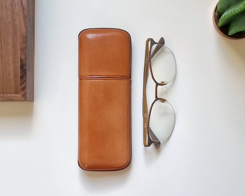 May include: A brown leather glasses case with a gold metal frame glasses case with clear lenses.
