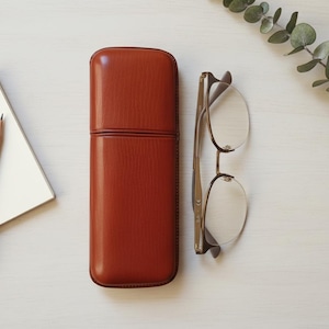 Puede incluir: Vista superior de una composición de escritorio con un estuche de gafas de cuero marrón rojizo cerrado, gafas, un bloc de notas, un lápiz y una ramita de vegetación. Las gafas tienen una montura fina de color dorado.