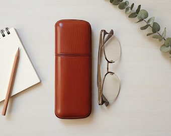 May include: A top-down view of a desk setup featuring a closed, reddish-brown leather glasses case, eyeglasses, a notepad, a pencil, and a sprig of greenery. The eyeglasses have a thin, gold-colored frame.