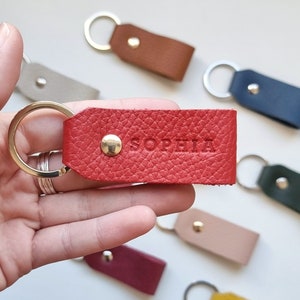 Può includere: Un portachiavi in ​​pelle rossa con un anello dorato e il nome "Sophia" inciso su di esso. Il portachiavi è tenuto in una mano, e ci sono altri portachiavi sullo sfondo.