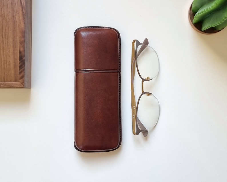 May include: A brown leather glasses case with a stitched seam and a pair of gold-rimmed glasses with clear lenses.