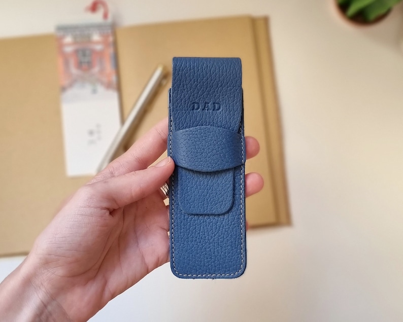 Leather pen case, it closes with a flap and the DAD monogram is engraved on the top. This pen holder is blue. It is shown close up to appreciate the high quality full grain leather with which it is handmade.