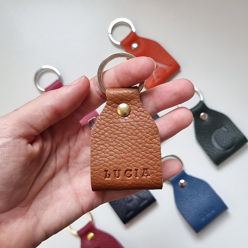 In one hand is a small leather keychain engraved with five initials (name of Lucia). Brown color, full grain leather. The keychain is on one hand and behind it you can see more keychains of other colors.