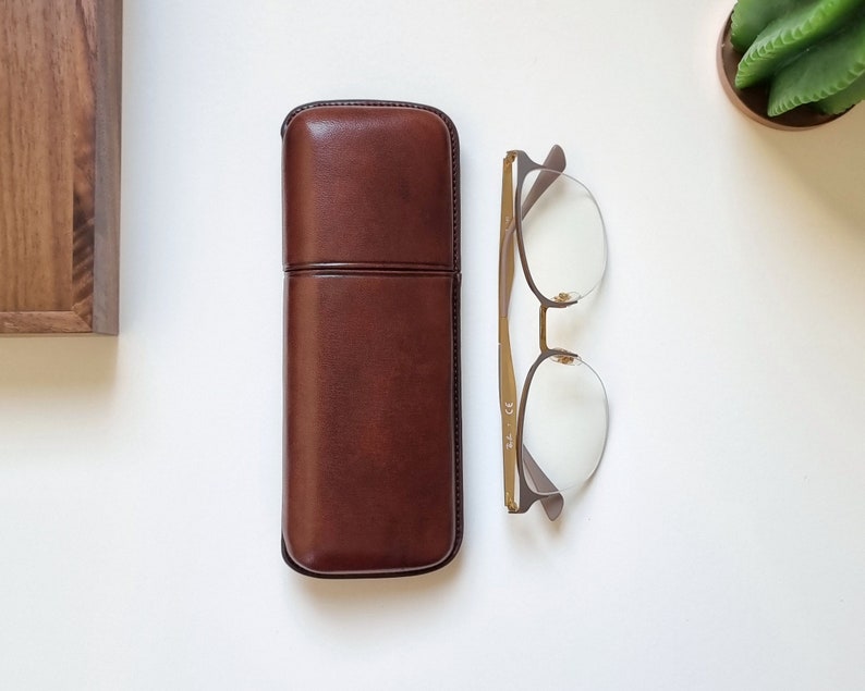 brown leather eyeglasses case in  on a white table. Shown closed with eyeglasses next to it to show the proportion between the eyeglasses and the leather case