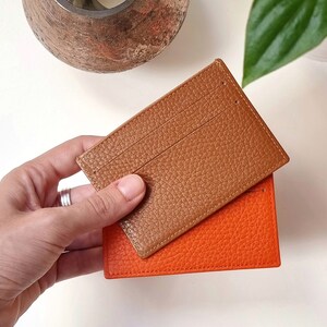 The leather used for these cardholders is very soft, high quality full grain leather. This card holder has two card slots. In a hand are shown two plain color cardholders, one in orange and the other in havana or brown.