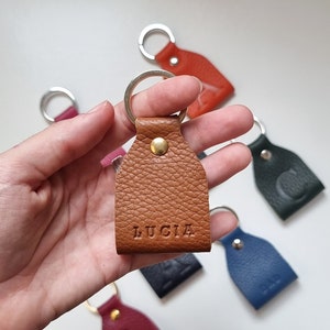 In one hand is a small leather keychain engraved with five initials (name of Lucia). Brown color, full grain leather. The keychain is on one hand and behind it you can see more keychains of other colors.